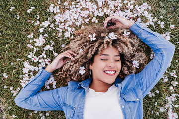 top view of happy hispanic woman with afro hair lying on grass among pink blossom flowers.Springtime - 494302481