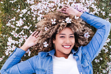 portrait of happy hispanic woman with afro hair lying on grass among pink blossom flowers.Springtime