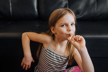 A beautiful, red-haired child with a sweet tooth sits on a black leather sofa with a lollipop in...