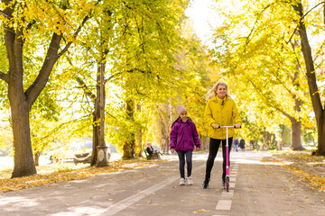 Fototapeta premium Mother and daughter are walking in the autumn in the park. woman is riding a scooter