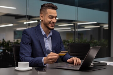 Fototapeta na wymiar Cheerful middle eastern entrepreneur using laptop, holding credit card