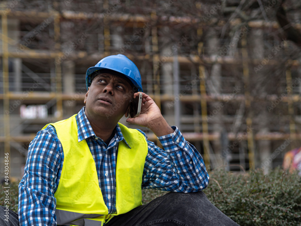 Wall mural Indian or Somali engineer with safety jacket and helmet talking on mobile phone on construction site