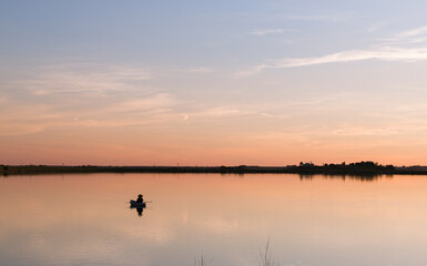 Fisherman floating on the still lake during sunset
