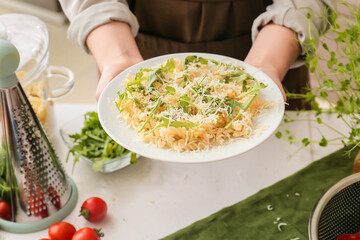 Woman holding plate of tasty pasta with arugula and cheese in kitchen, closeup