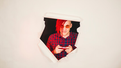 Young man in red plaid shirt with gamepad and playing video game. Impressive hipster in eyeglasses with red dreadlocks gamer playing computer games through a hole in the white wall in the studio