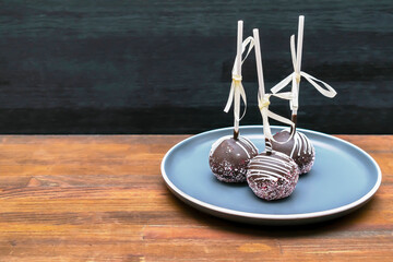 Delicious sweet chocolate cake pops, popsicles decorated white sprinkles on plate, wooden background, pastry, desserts on stick with tape. Tasty food. mock up, template, copy space
