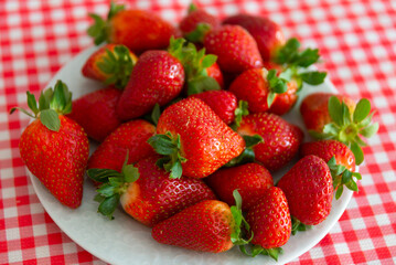 Fresh strawberries with green leaves in a white plate on a red and white tablecloth.