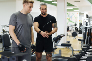 Fototapeta na wymiar Fit man discussing workout plan with his trainer, personal coach before starting workout in the gym. Personal coach standing near young man and showing something on his clipboard