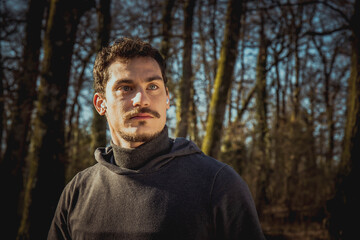 Runner is listening to the music while he stretches in the woods. Portrait of a handsome man with blue eyes and a mustache who is training in the park.