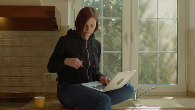 Young adult woman working from home using laptop sitting in the kitchen and drinking coffee.