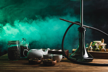 Hookah, dry tea and teapot on wooden surface on black background with smoke.