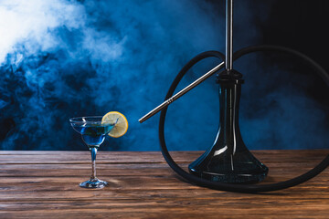 Glass with cocktail near hookah on wooden surface on black background with smoke. © LIGHTFIELD STUDIOS