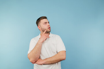 Pensive man in a white T-shirt and with bristles stands on a blue background with a serious face, looks away and thinks
