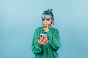 Funny girl with colored hair and smartphone in her hands looks away with a pensive face isolated on blue background