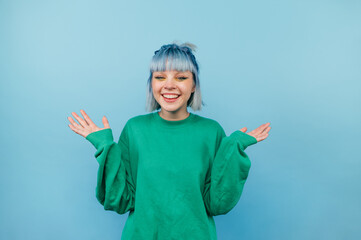 Positive teen girl with blue hair and in a green sweater rejoices with raised hands on a colored...