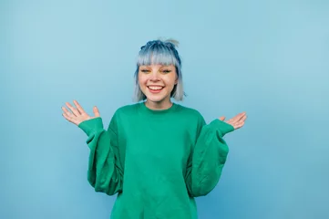 Fotobehang Positive teen girl with blue hair and in a green sweater rejoices with raised hands on a colored background. © bodnarphoto