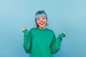 Joyful girl with blue hair in a green sweatshirt rejoices with a smile on his face and closed eyes on a blue background