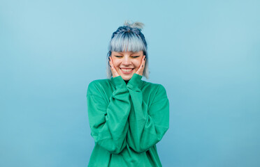 Joyful lady in green sweatshirt and blue hair is happy with a smile on her face on a blue background.