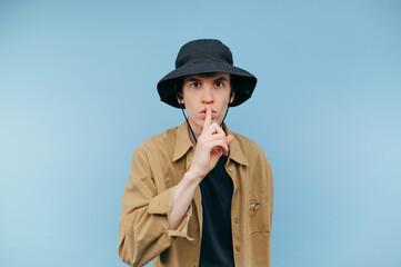 Handsome hipster guy in panama and shirt stands on a blue background, shows a gesture of silence in the camera with a serious face
