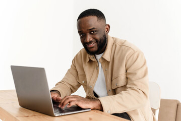 Portrait of smiling african american man looking at camera while sitting at table in home office and working on laptop. Happy black freelancer posing at workplace at home.