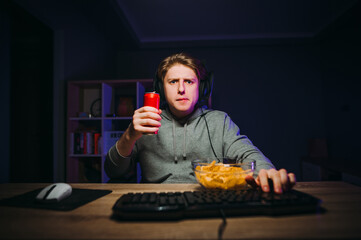 Serious guy in casual clothes sits on the computer in a headset with a drink in his hands and a...