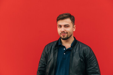 Handsome adult man in a leather jacket and with a beard stands on a background of a red wall and looks at the camera with a smile on his face.