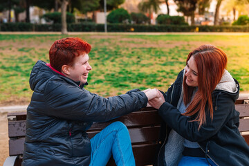 Fototapeta na wymiar Couple of two lesbian women, smiling, sitting, holding hands on a bench in a public park.
