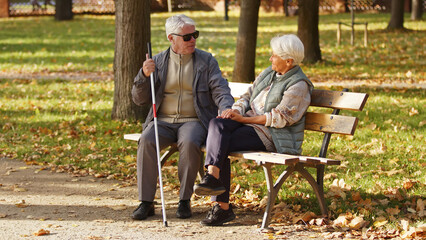 Elderly Caucasian couple sitting on the bench looking at each other in a park woman holds her husbands' hands disabled blind man with glasses and a stick blind people emotional support rehabilitation
