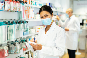 Oriental woman doctor in face mask standing in chemist shop with recipe blanks and pen in hands.