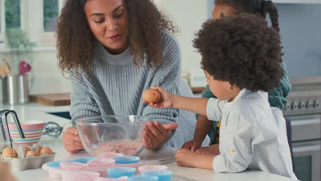 Mother and children in kitchen breaking egg into bowl and mixing ingredients together for cupcakes - shot in slow motion