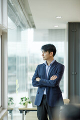 Portrait of young happy Asianbusinessman wearing grey suit and blue shirt standing in his office and smiling with arms crossed