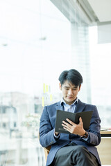 Portrait of young happy Asianbusinessman wearing grey suit and blue shirt standing in his office and smiling with arms crossed