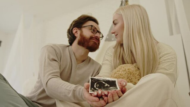 soon-to-be caucasian parents sitting on the floor and showing to the camera an ultrasound scan of pregnancy. High quality 4k footage