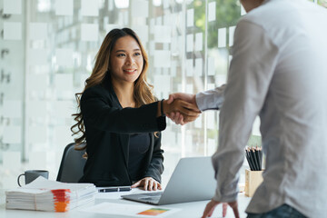 Smiling asian manager sitting at her desk in an office shaking hands with a job applicant after an...