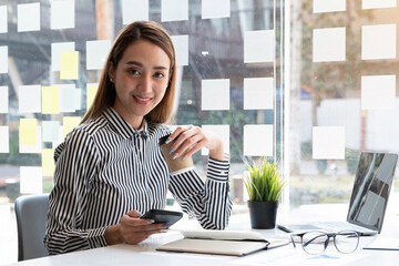 Beautiful young Asian businesswoman drinking a coffee and holding a smartphone working on laptop at office. Looking at camera.