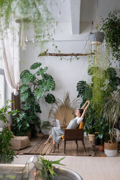 Happy relaxed woman remote worker resting, stretching arms while working at nature-inspired home office filled with many different exotic indoor plants, sitting with laptop on chair in home garden