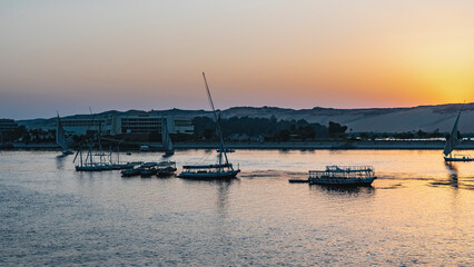 Fototapeta premium Evening Nile. Felucca and tourist boats on calm water. The sun hid behind a sand dune. The sky is highlighted in orange. Glare on the river. Egypt. Aswan