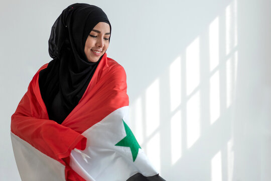 Happy Muslim woman wears black Hijab and Muslim traditional clothes wrapped in Irag flag, sunlight and shade on white wall, Muslim woman rights concept