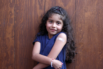 Happy and excited 7-year-old curly-haired little Latina girl shows her arm recently vaccinated...