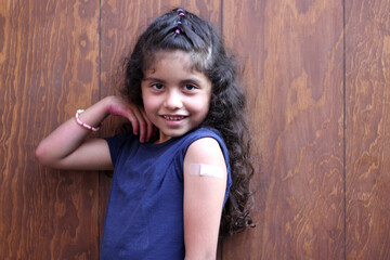 Fototapeta premium Happy and excited 7-year-old curly-haired little Latina girl shows her arm recently vaccinated against Covid-19 in the new normal for the coronoavirus pandemic 