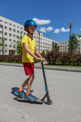 A young man in a yellow T-shirt and a blue helmet rides a stunt scooter through the city.