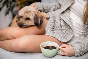 A woman sits on a bed with coffee in hand and a multiracial dog next to her.