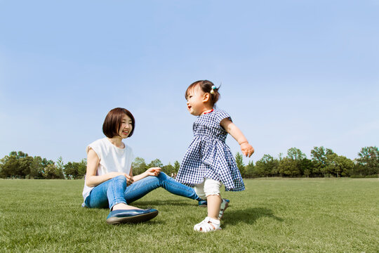 青空を背景に緑の芝に座り幼い娘と一緒に遊ぶ若い母。幸せ,愛情,母性,育児イメージ