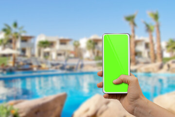 female hand holding modern mobile phone with blank green desktop screen on hotel resort background