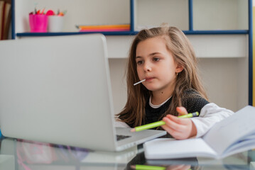 A happy kid student eating a lollipop while learning for school on a laptop at the home desk....