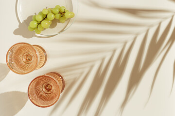 Wine glasses from color glass and green grape on transparent plate, beige background, palm leaf shadows from sunlight. Summer drinks concept. Trend wine glass goblets style, colored glassware