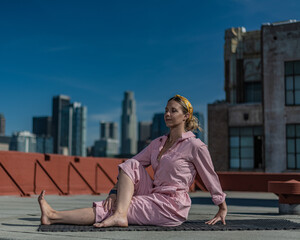 Young blonde woman in pink coveralls and barefoot practices yoga on a downtown Los Angeles Roof top with the LA skyline behind her