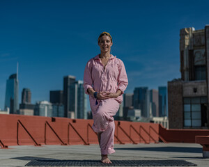 Young blonde woman in pink coveralls and barefoot practices yoga on a downtown Los Angeles Roof top with the LA skyline behind her