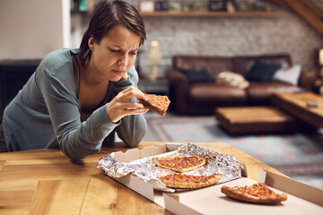 Young unhappy woman eats pizza due to her depression at home.