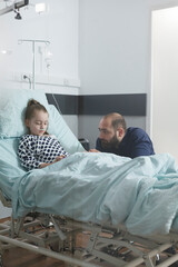 Anxious sad man watching unwell little daughter sleeping in clinic bed while in pediatric hospital patient ward. Under treatment sedated ill child resting while attentive parent taking care of her.